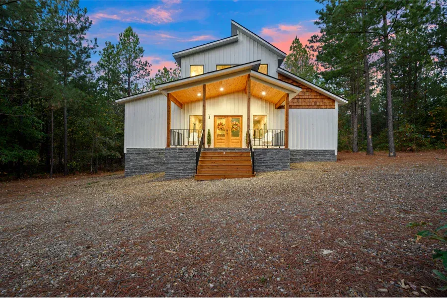 A modern white house with a stone base and wooden front porch sits at the edge of a forest under a sunset sky.