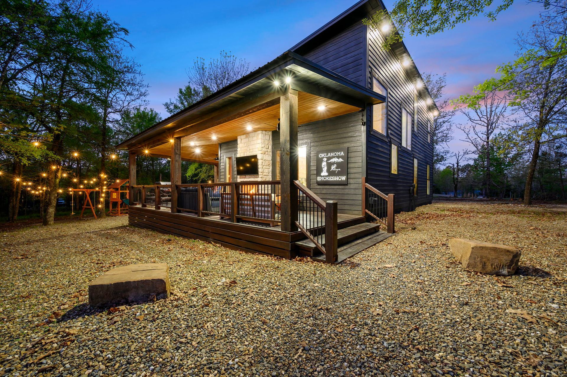 A dark wooden cabin with a lit-up porch and outdoor fireplace sits on a gravel lot at twilight with string lights nearby.