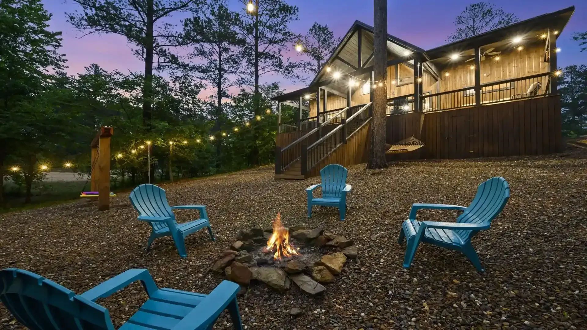A fire pit with four blue chairs in front of a rustic wooden cabin at dusk, illuminated by string lights.