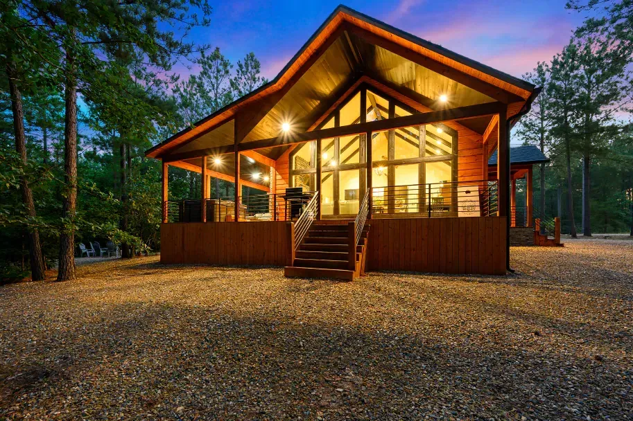 A modern wooden cabin with large glass windows and a front deck illuminated at twilight in a wooded area.