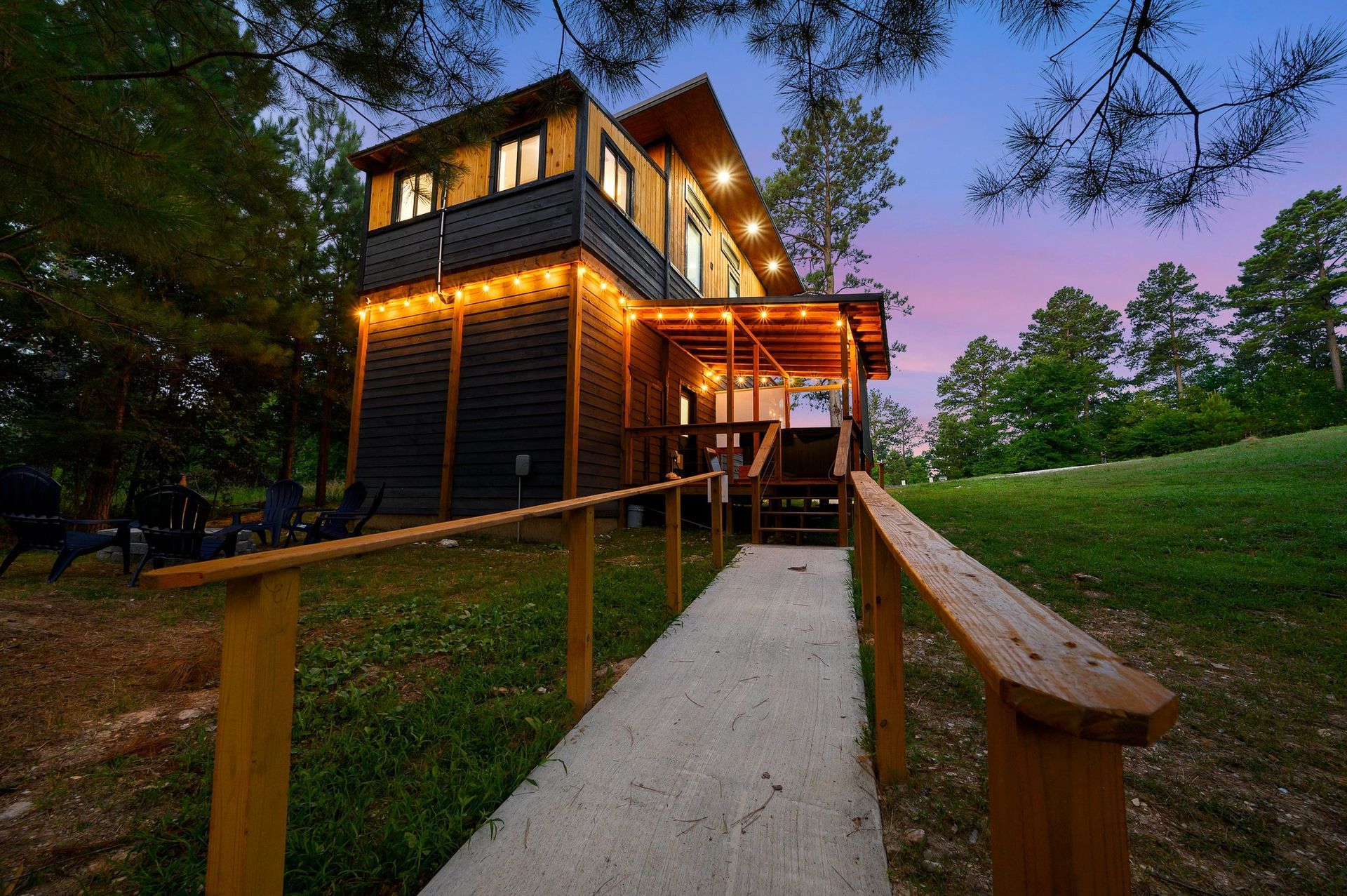 A modern, two-story wooden cabin at dusk, illuminated by warm string lights, with a concrete ramp leading to the entrance.
