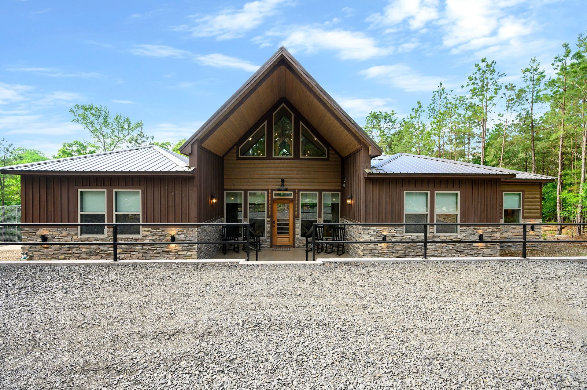 Wooden lodge with a steep roof and stone accents, centered behind a gravel lot under a blue sky.