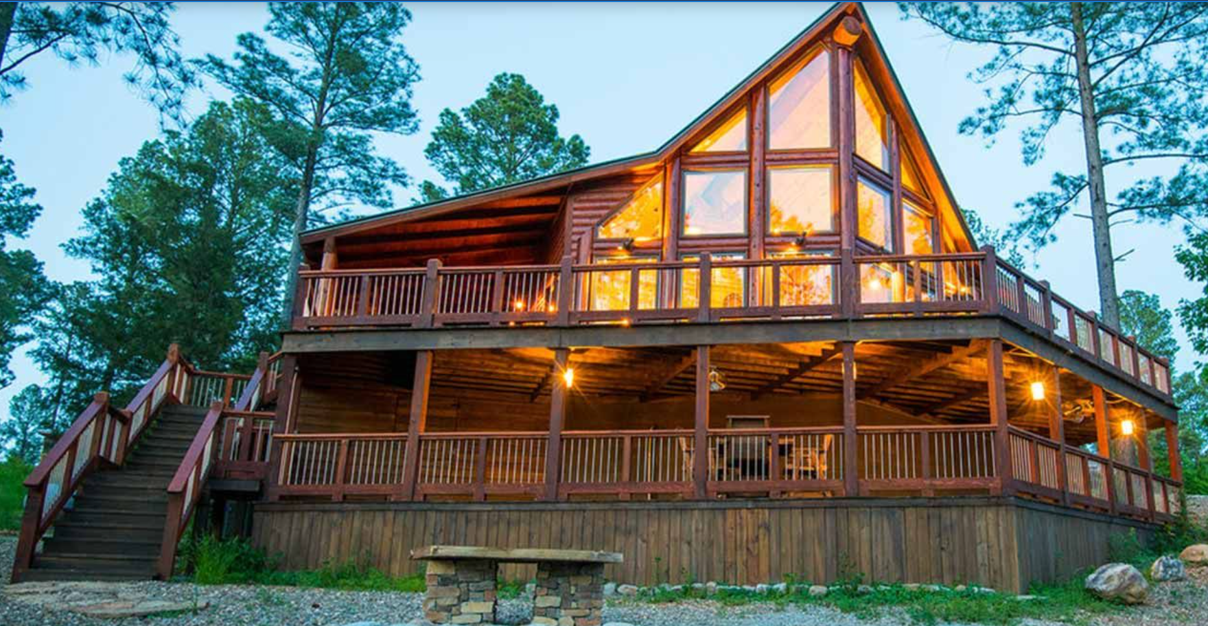 A two-story rustic wooden cabin with a large deck and expansive windows glowing with warm light, set in a pine forest.