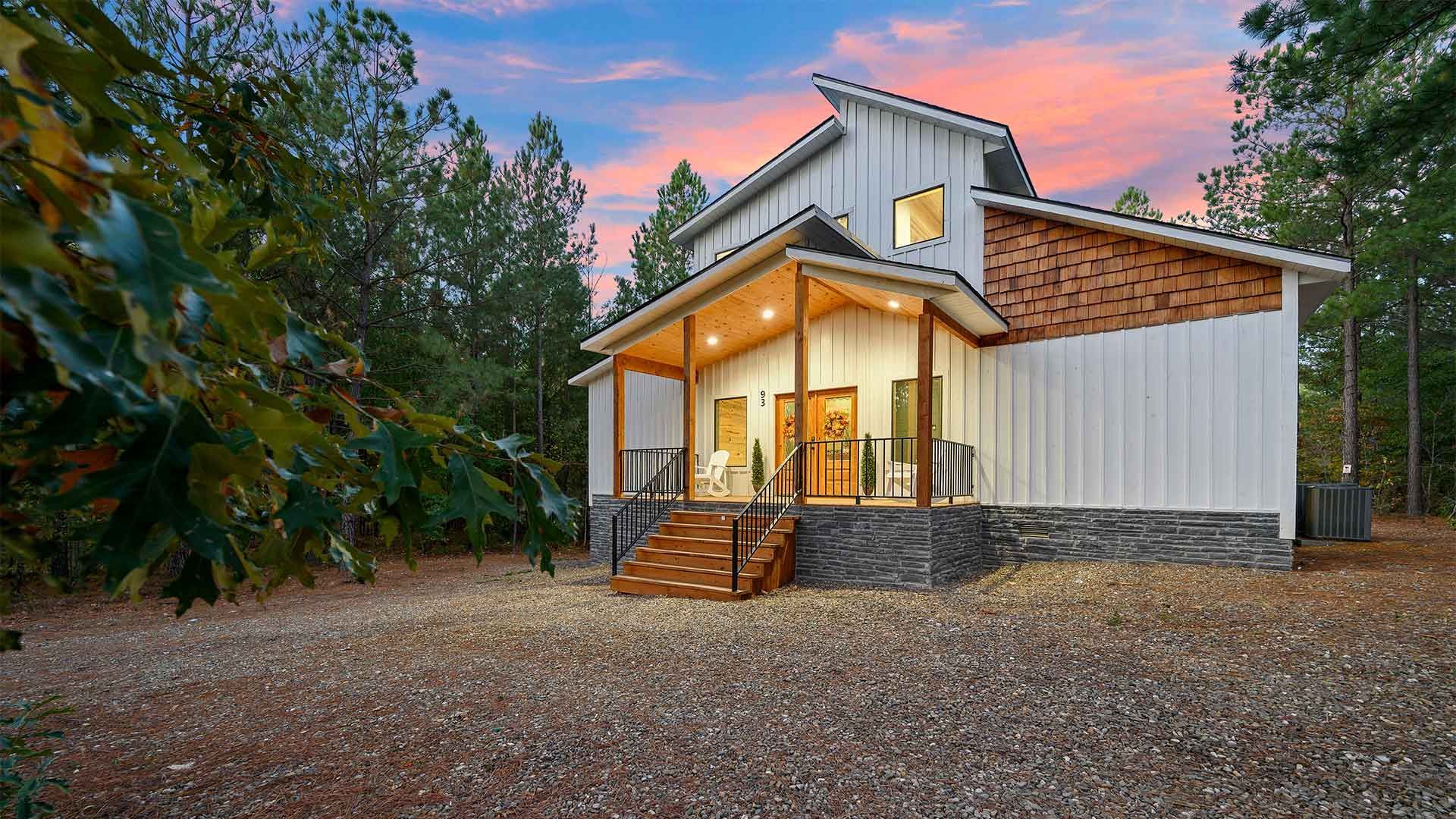 A modern white farmhouse with a stone foundation, wood accents, and a covered porch, set among trees at sunset.