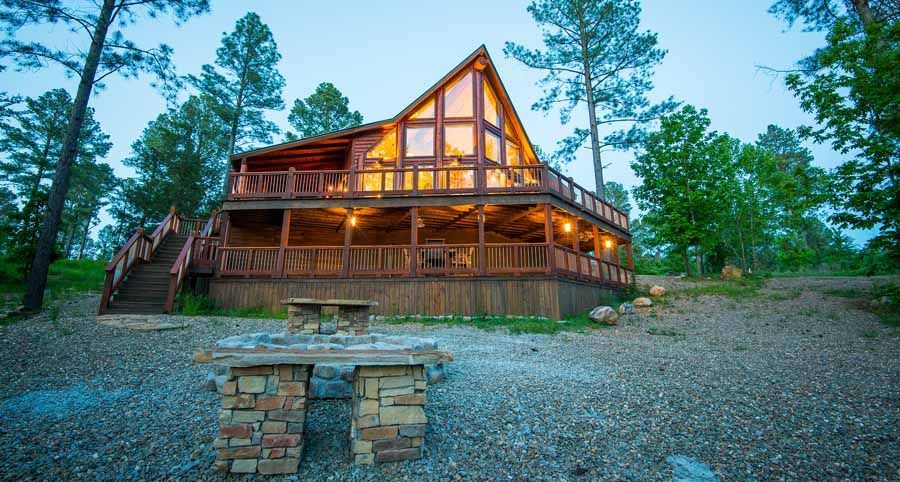 A rustic, multi-story wooden cabin illuminated at twilight, surrounded by trees with a stone fire pit in the foreground.