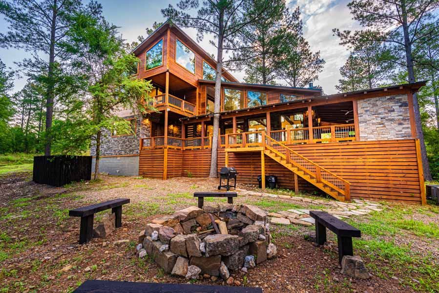 A three-story wood cabin with stone accents and a large deck sits in a pine forest near a stone fire pit with benches.