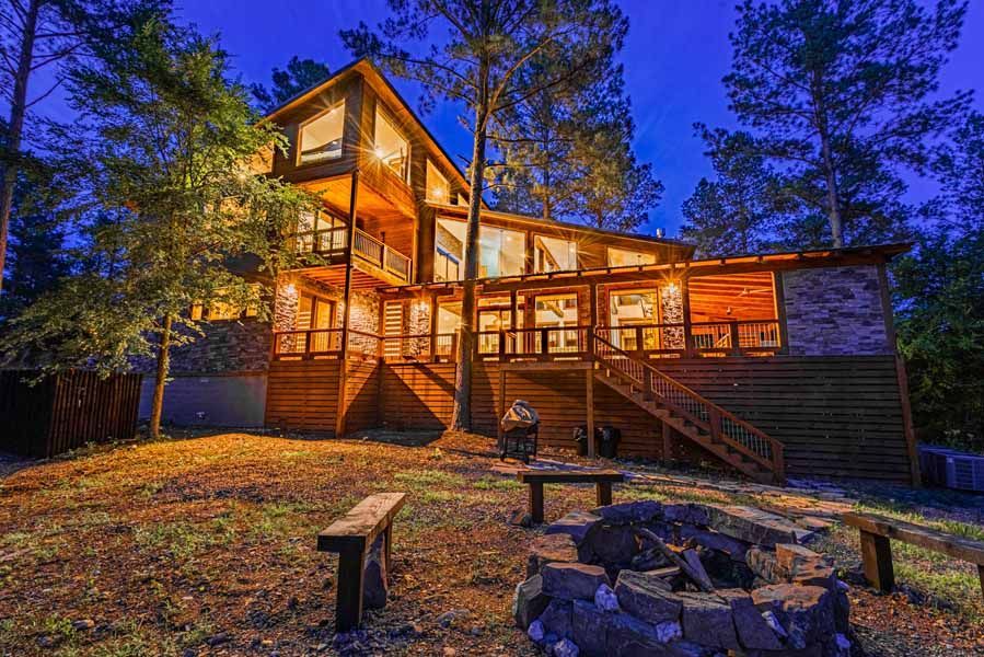 A multi-story wooden cabin illuminated at night, featuring a large deck and a stone fire pit in the foreground.