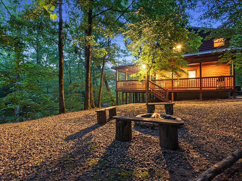A cabin with a glowing, lit porch set in a forest at dusk, featuring a gravel area with a fire pit and wooden benches.