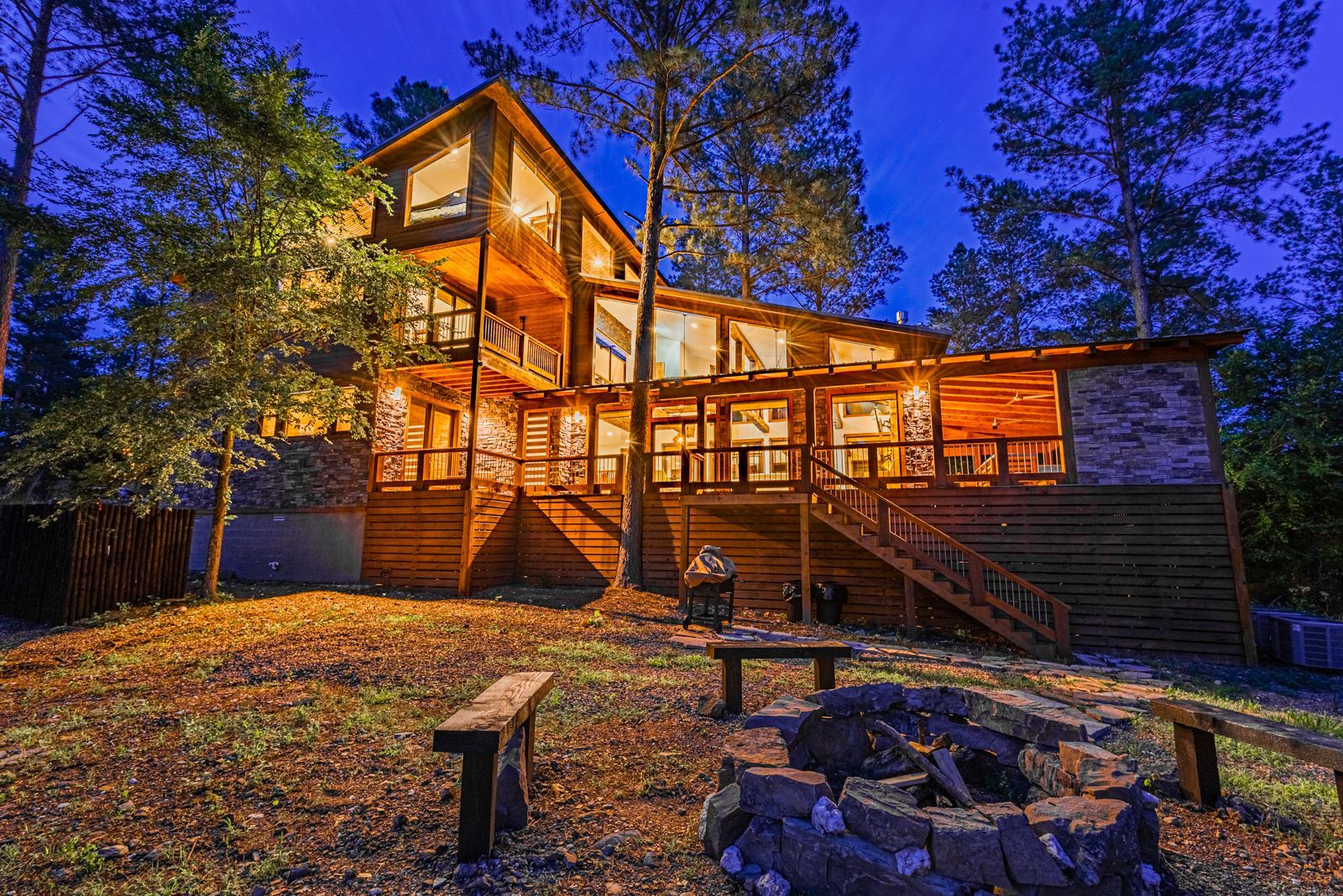 A large, multi-level wooden cabin illuminated at night, featuring a wraparound deck and a fire pit in the foreground.
