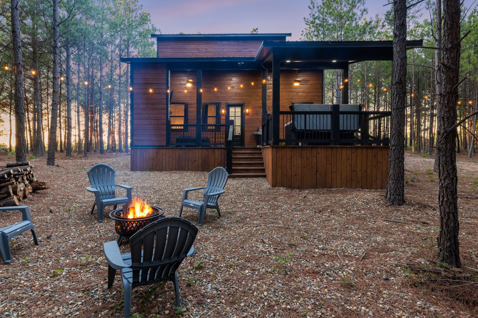 A rustic A-frame cabin with a red metal roof sits among lush green trees at sunset with a gravel driveway in front.