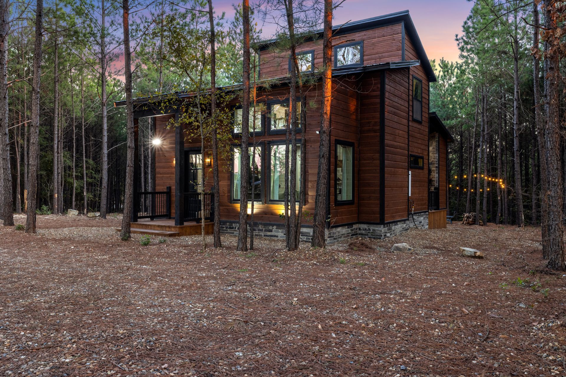 Modern cabin nestled among tall trees at dusk, with warm lights glowing through large windows.