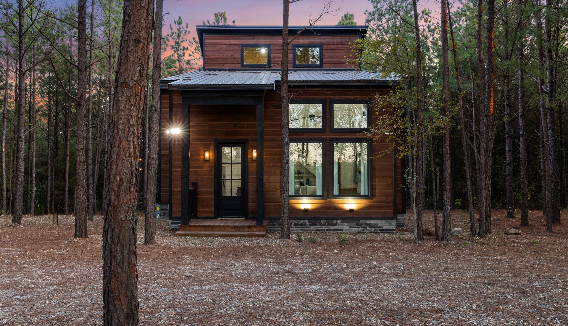 Modern two-story cabin with lit windows among tall pine trees at dusk
