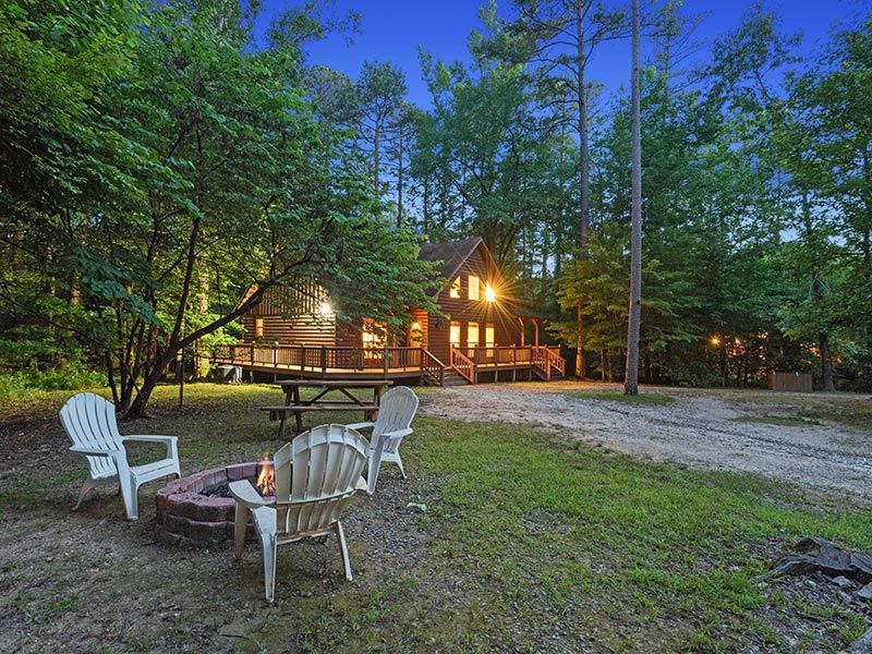 A cozy log cabin at dusk, nestled in a forest with a fire pit, Adirondack chairs, and a wooden picnic table.