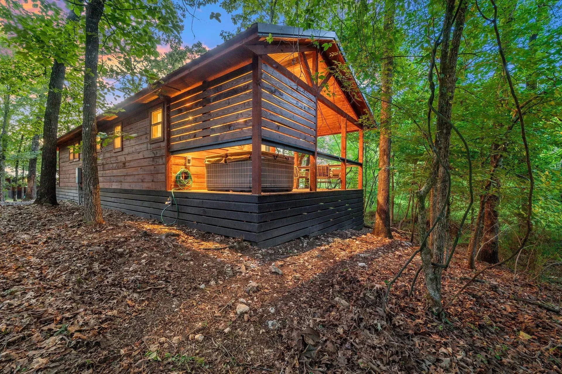 A cozy wooden cabin tucked in a dense, green forest at dusk, featuring a private porch with a hot tub.