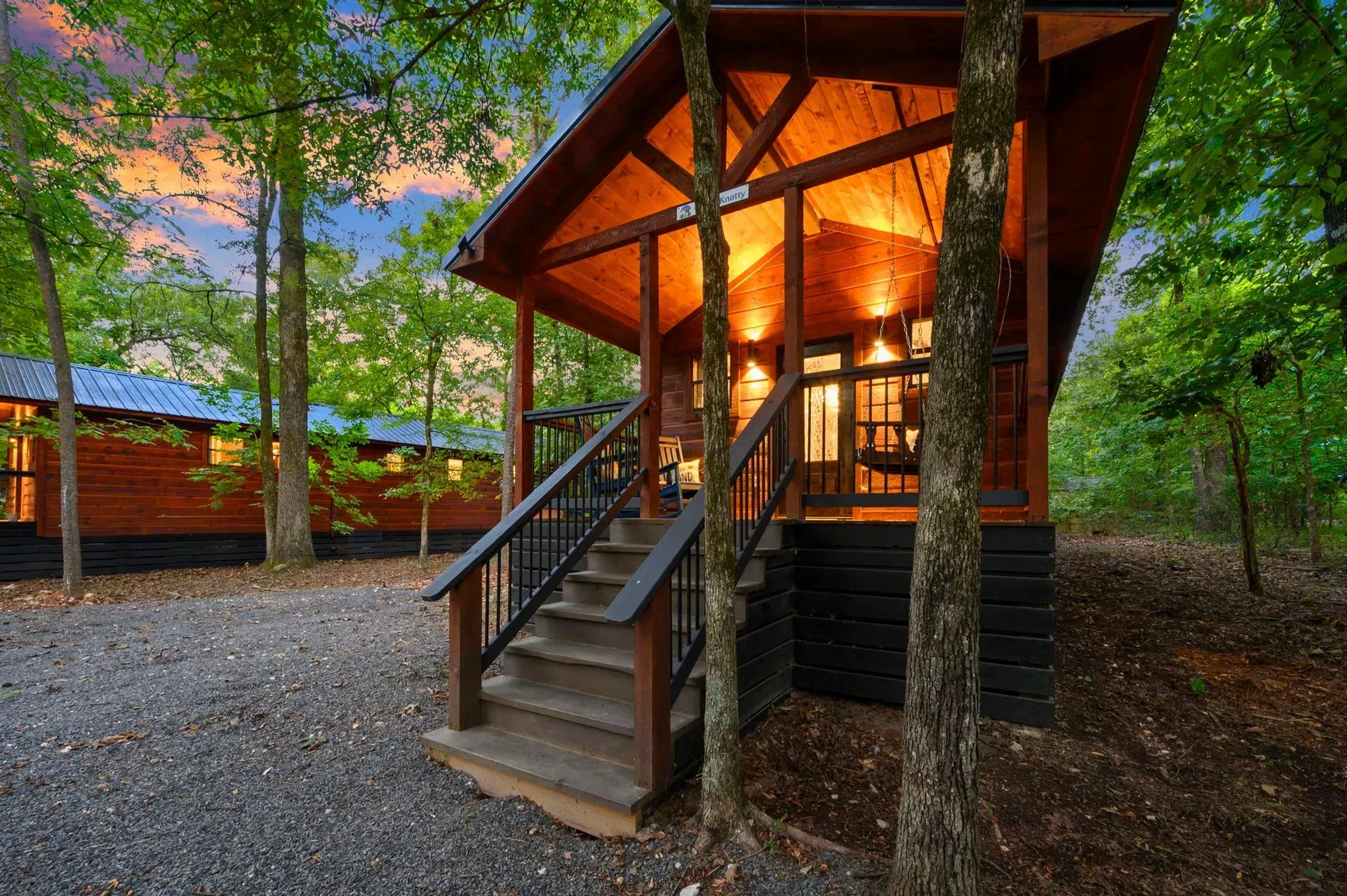 A rustic wooden cabin with a lit porch and stairs, nestled in a lush, green forest at sunset.
