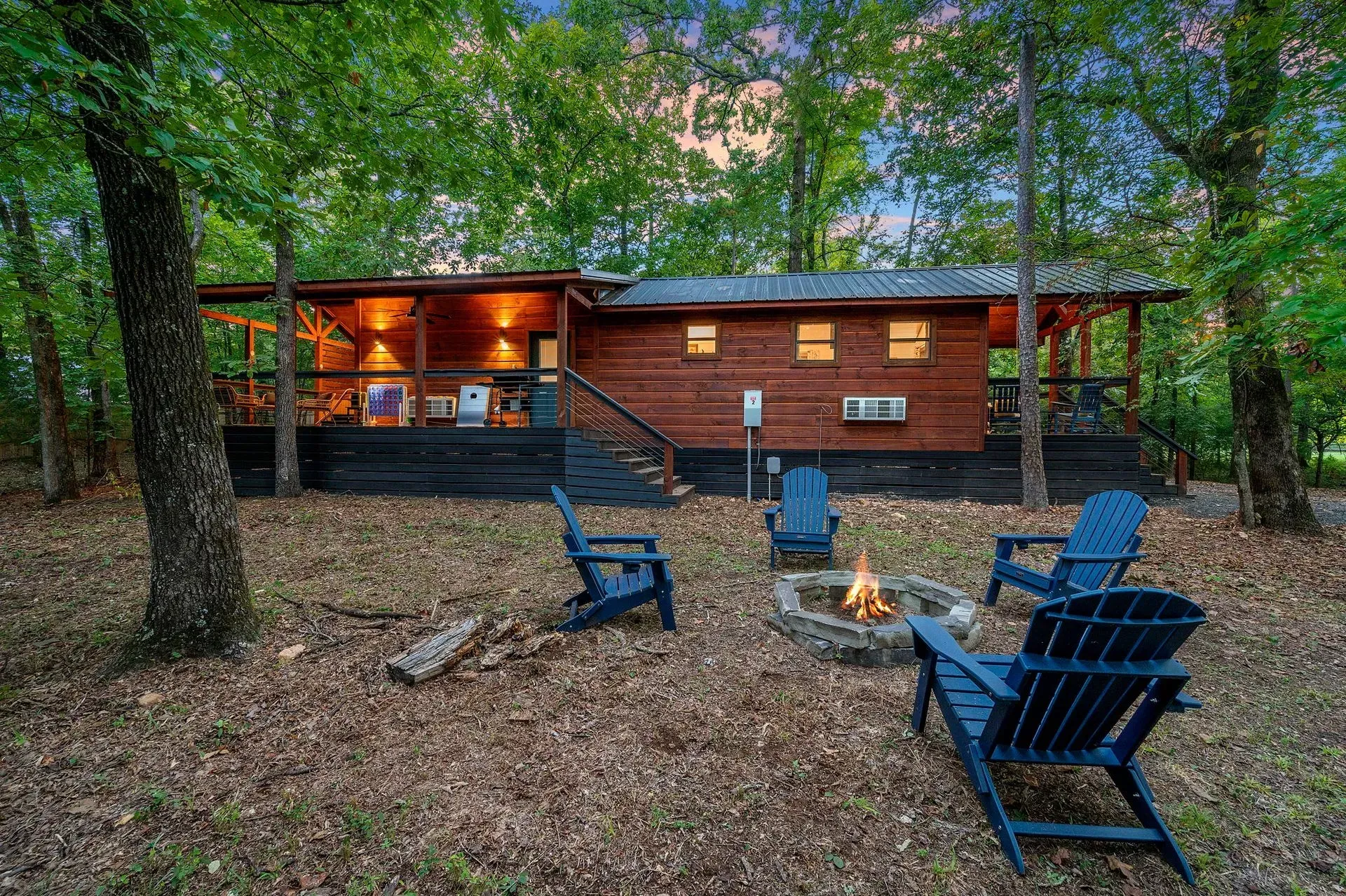 A rustic brown cabin with a wrap-around porch in a wooded setting, featuring a fire pit surrounded by blue chairs.