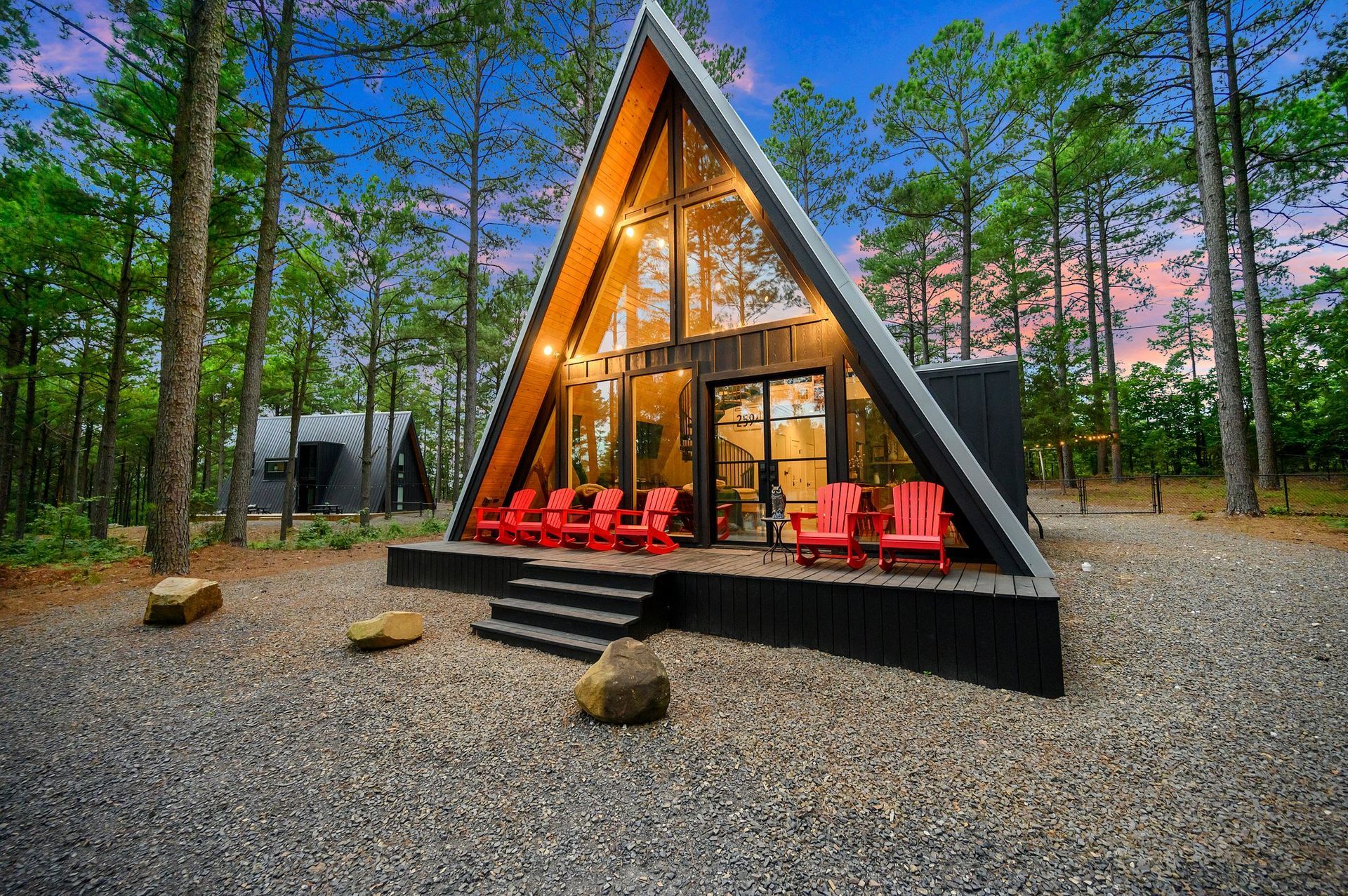 A modern, dark-framed A-frame cabin with floor-to-ceiling windows and red patio chairs, set in a forest at dusk.