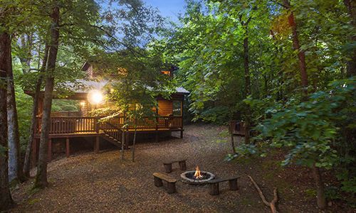 A cabin in the woods with a fire pit in front of it.