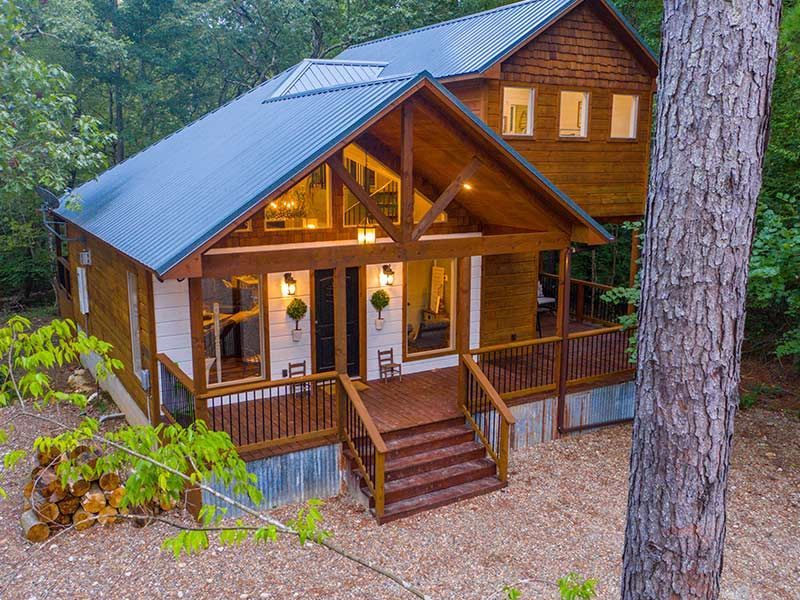 A two-story cabin with a metal roof, wooden siding, and front deck sits in a wooded area with a woodpile to the side.
