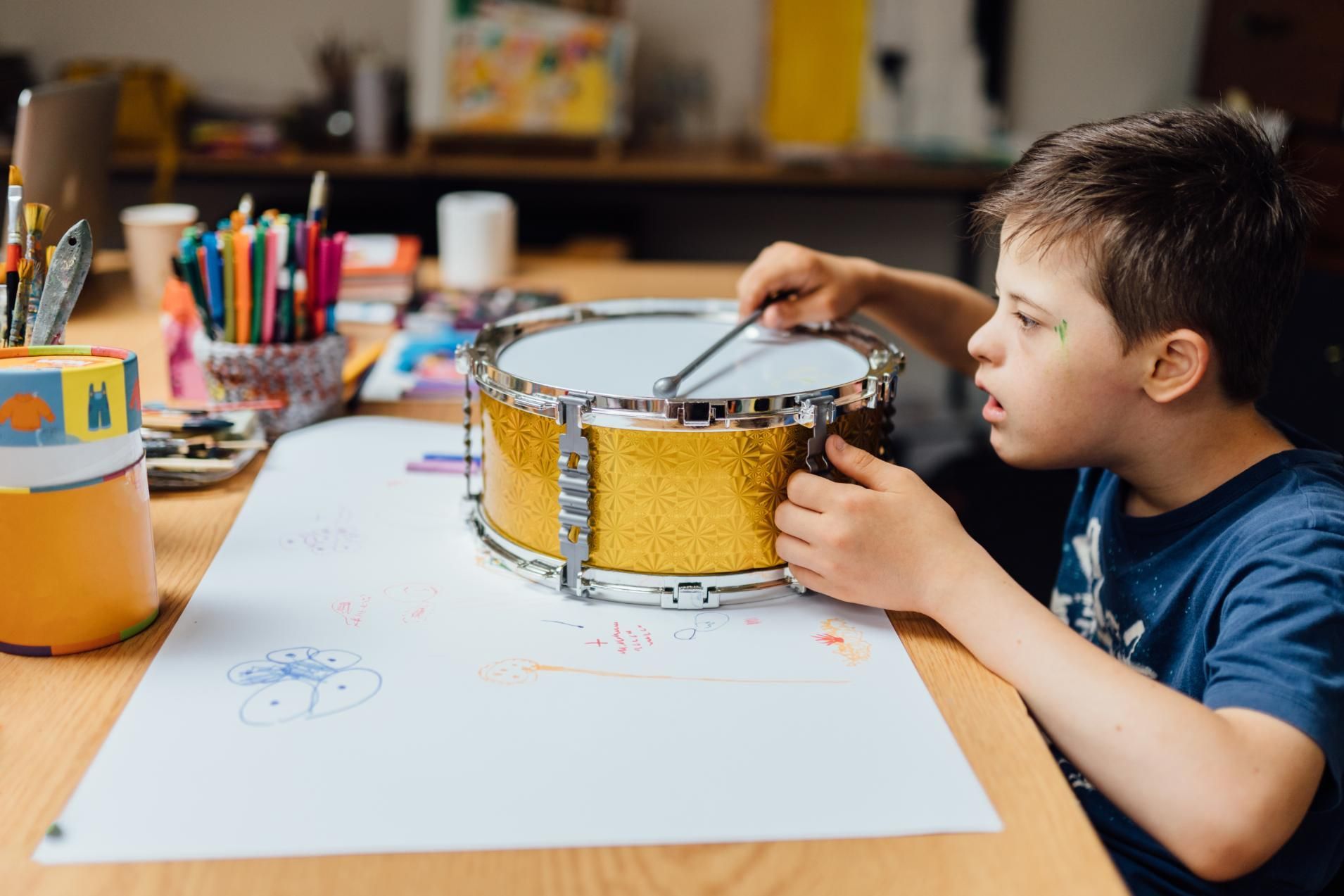 Child playing a yellow drum at a table with art supplies and drawings nearby