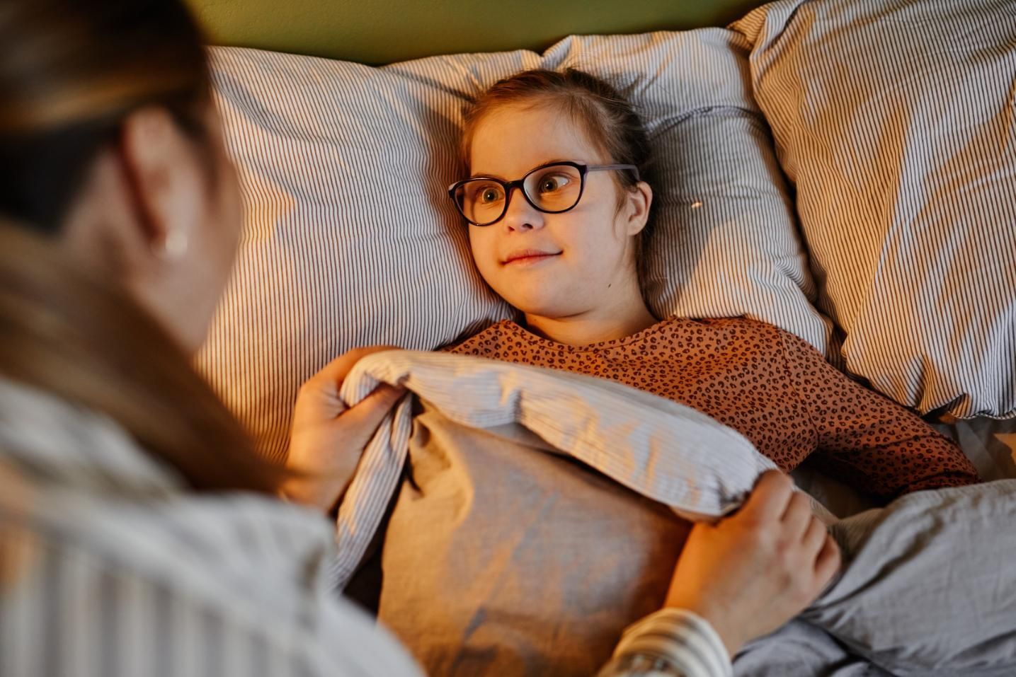 Child in glasses tucked under a blanket in bed, looking at an adult beside them.