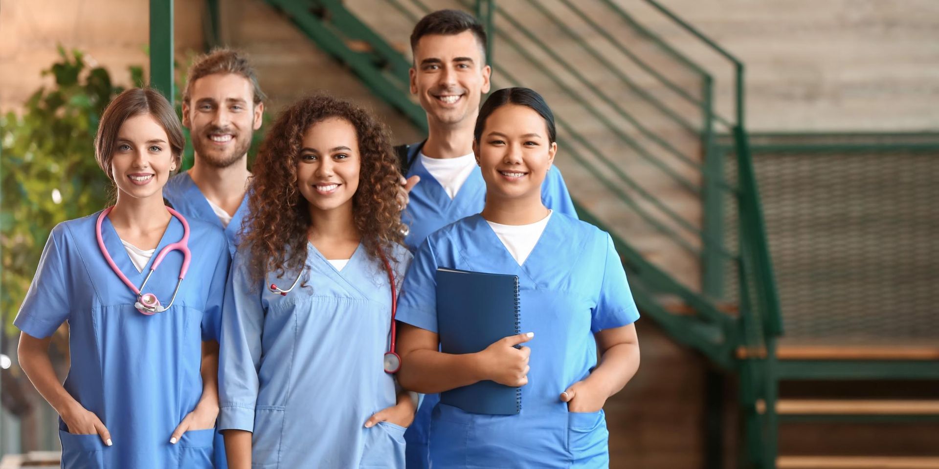 Five healthcare workers in blue scrubs standing together on hospital steps, smiling at the camera