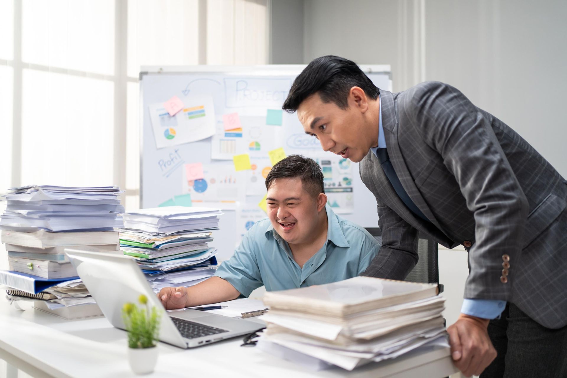 Two coworkers reviewing documents and a laptop in a bright office with stacks of paperwork