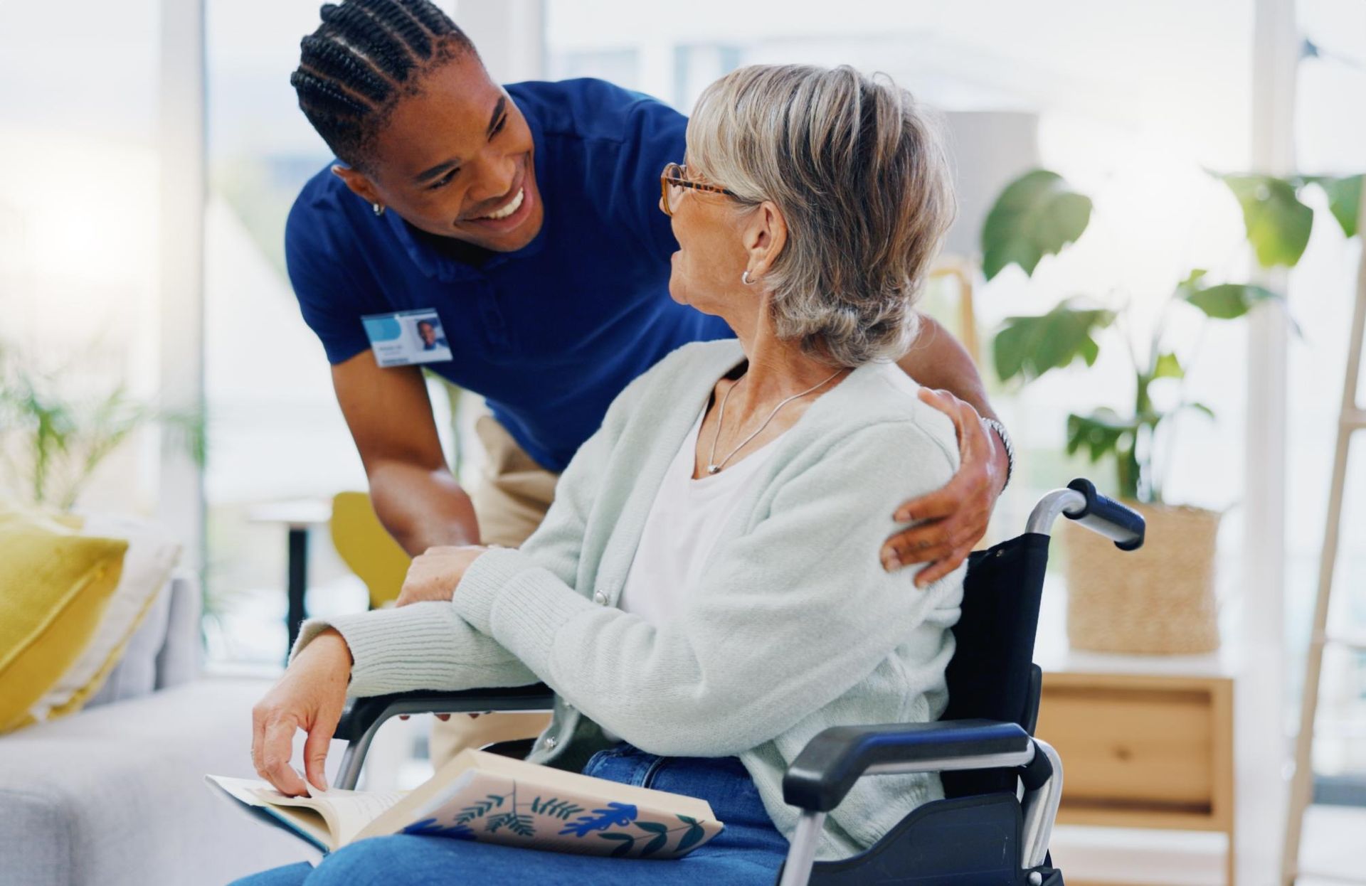 Caregiver in blue uniform smiling and talking with a seated person in a wheelchair at home