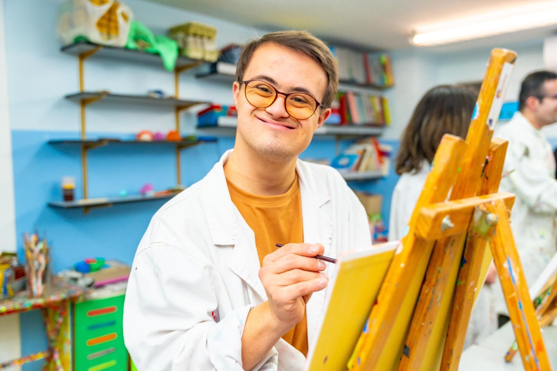 Person painting on an easel in a bright art studio, wearing glasses and a white coat.