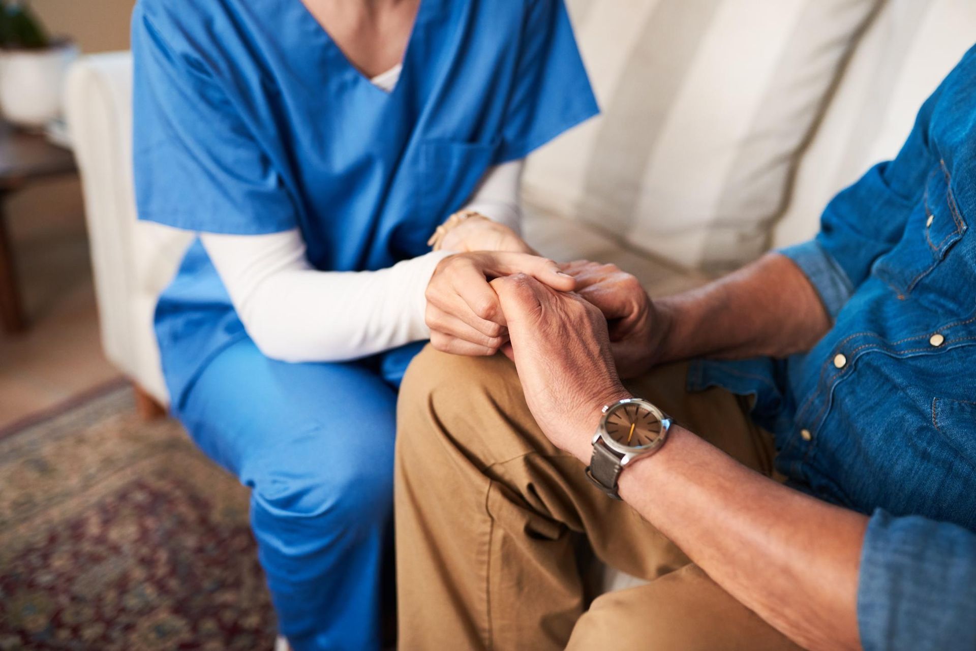 Caregiver in blue scrubs holding the hand of a seated person in a denim shirt, offering comfort