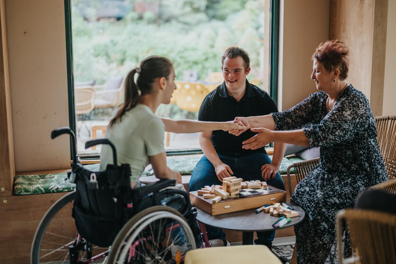 Three people sharing tea and snacks at a table, with one person in a wheelchair, by a window