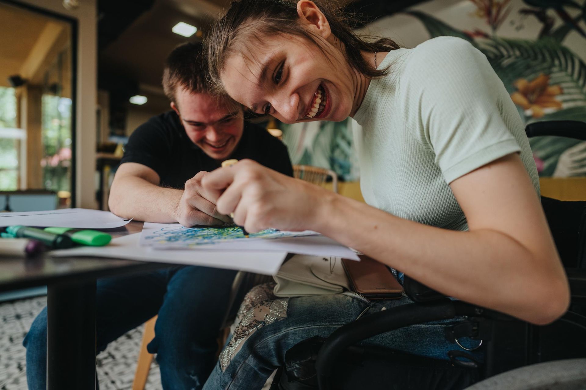 Two people sketching together at a table, one smiling over the paper in a bright room