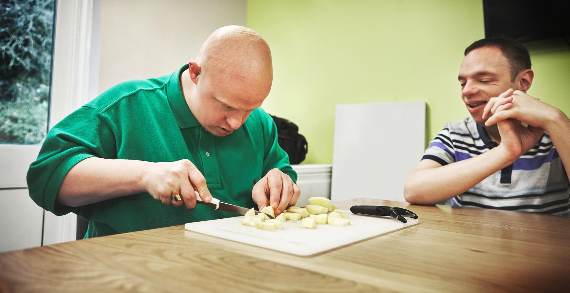 Two people at a table, one cutting food on a tray while the other watches and smiles