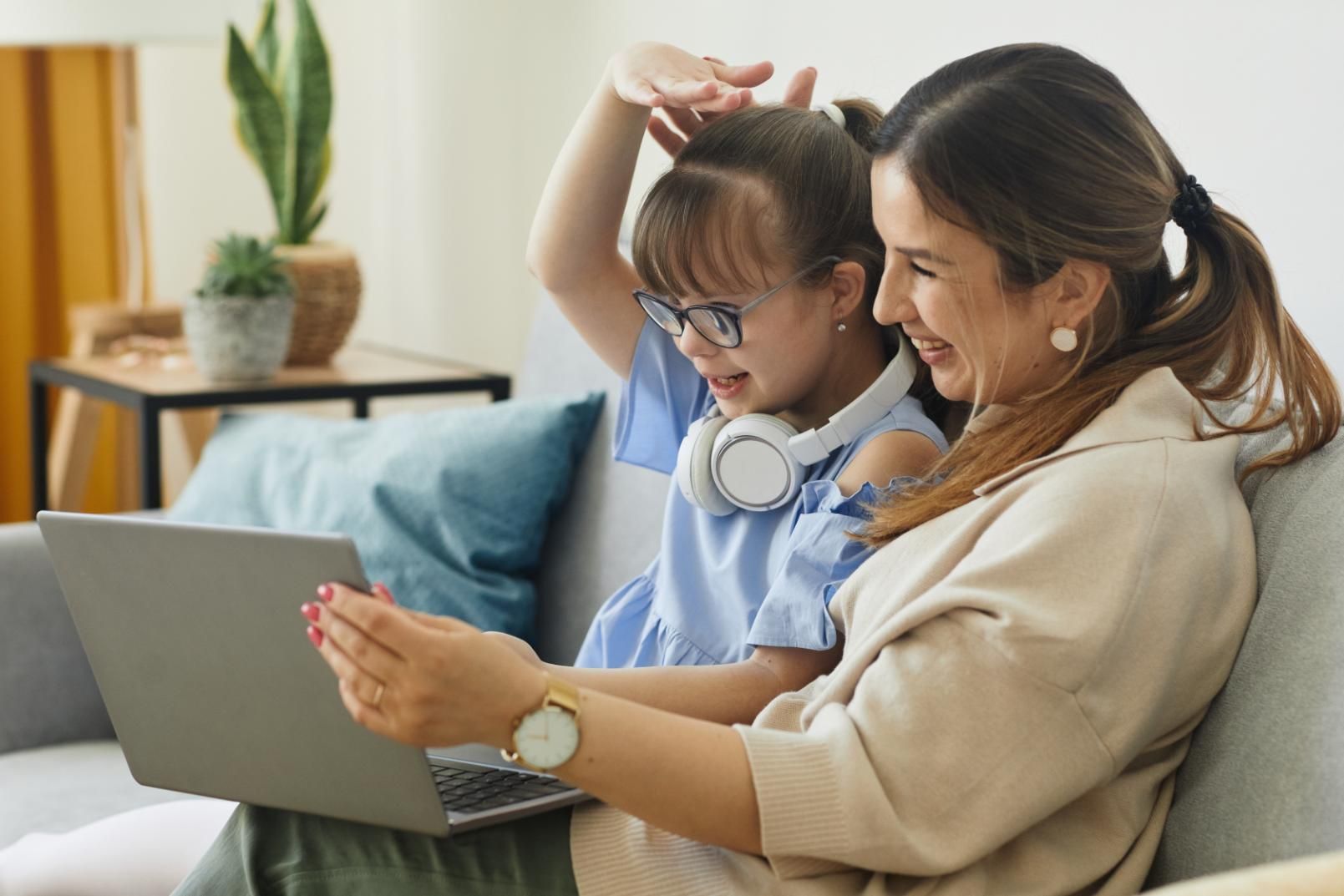 Woman and child using a laptop on a couch, with the child wearing headphones and raising a hand