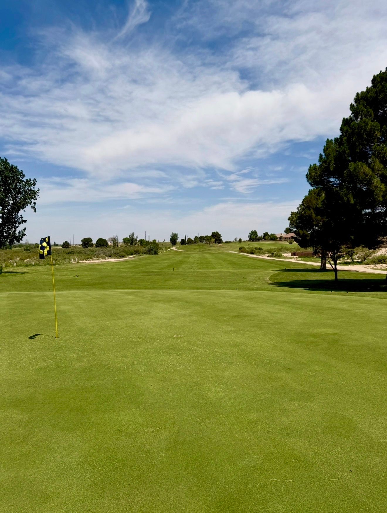 Green golf course with pond, trees, and buildings under a blue sky.