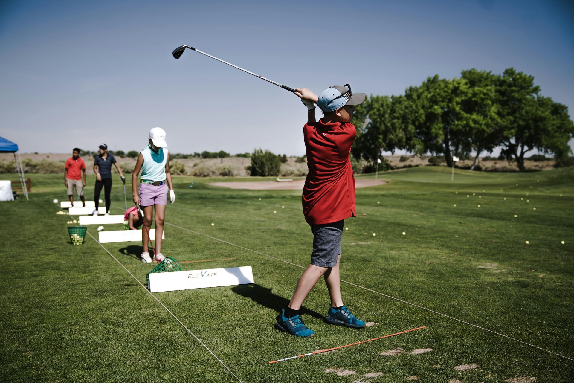 Person in red shirt swings golf club on a sunny driving range. Others watch.