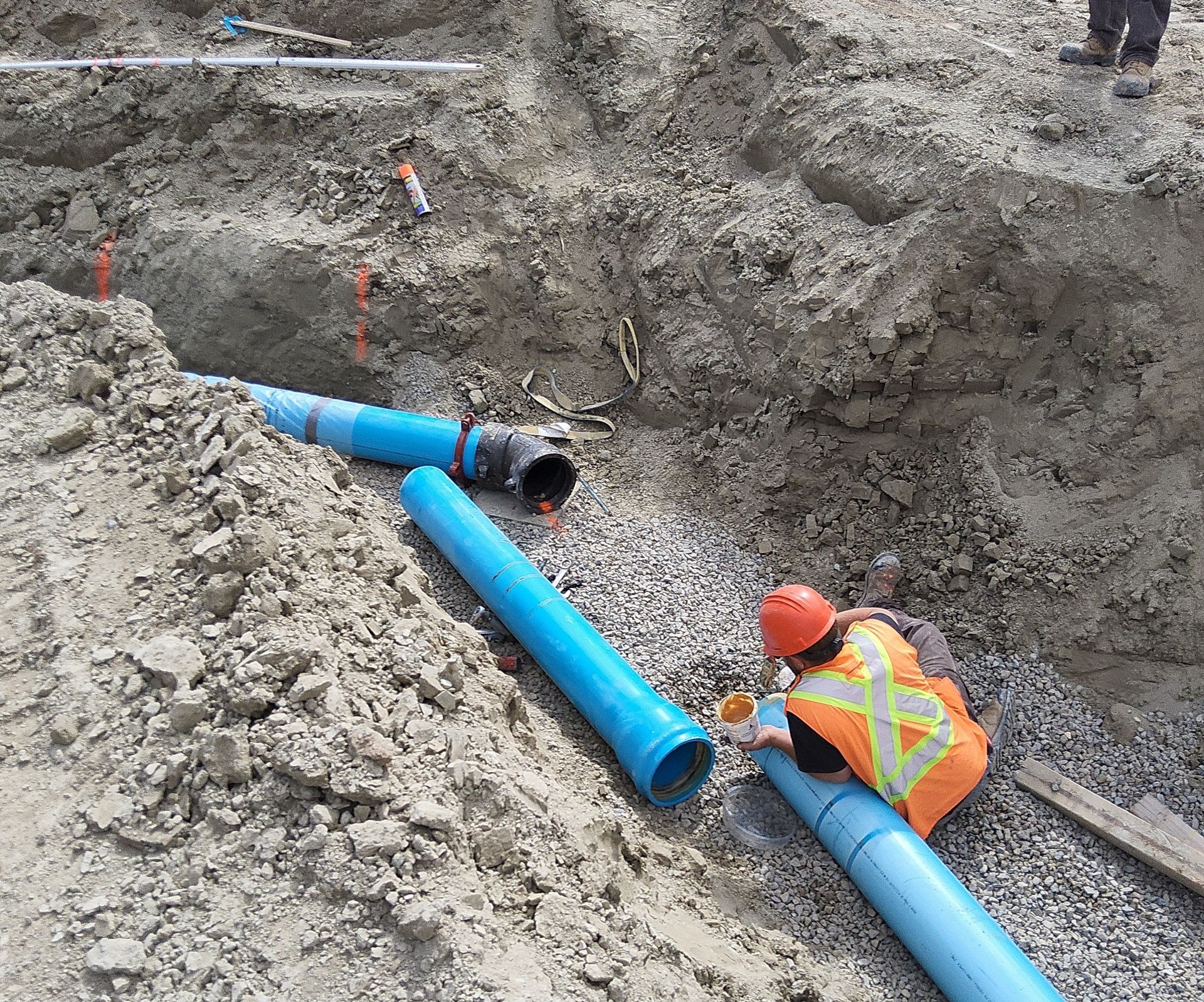 Worker in orange vest and hard hat installing blue pipes in a muddy trench at a construction site