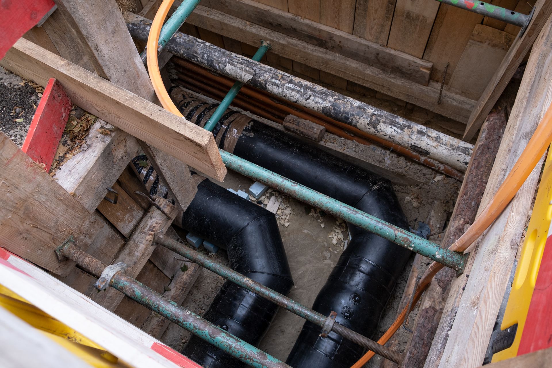 Construction pit with black pipes and green crossbars inside a wooden-framed trench
