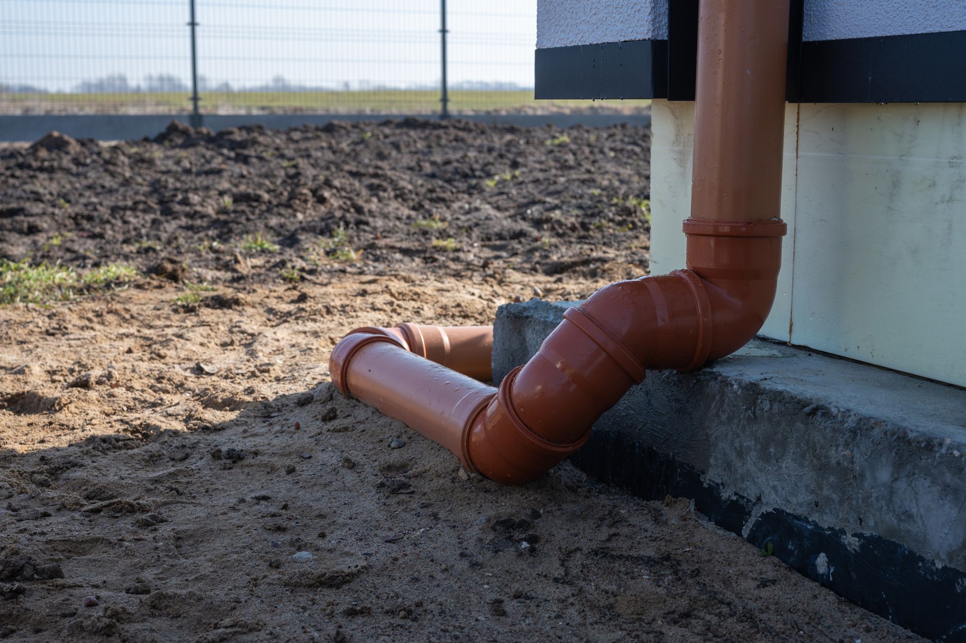 Orange PVC drain pipes connected to a building foundation on bare dirt.