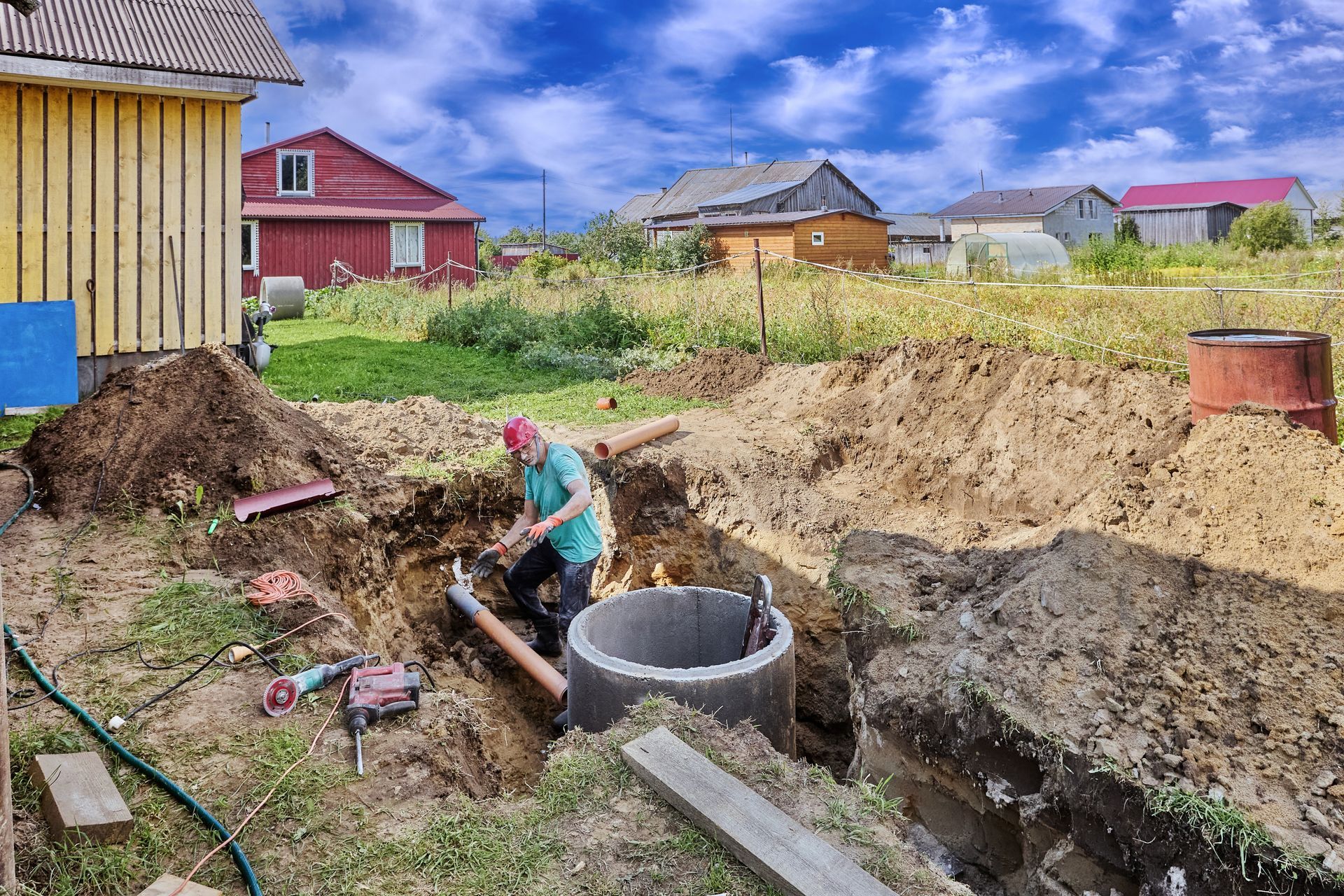 Worker digging a trench beside a circular concrete well in a rural yard with houses nearby