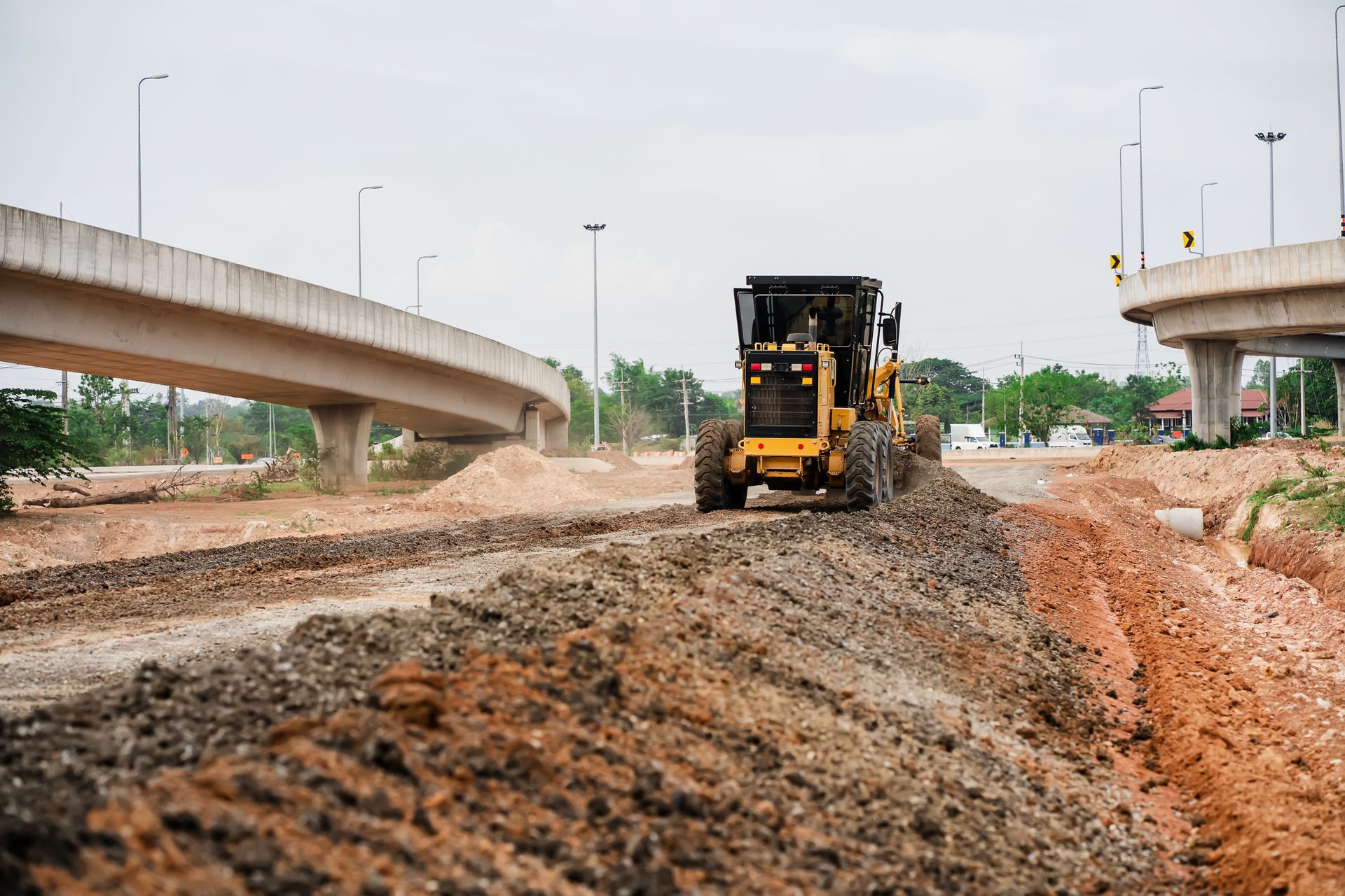 Construction machinery grading dirt beneath highway overpasses on a muddy road site