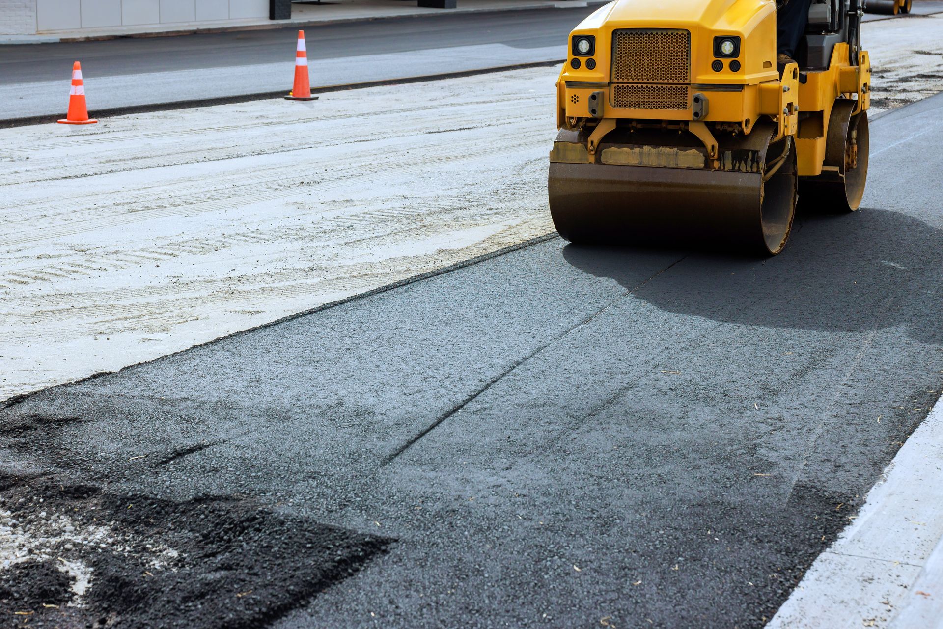 Yellow road roller paving a gray roadway beside orange traffic cones