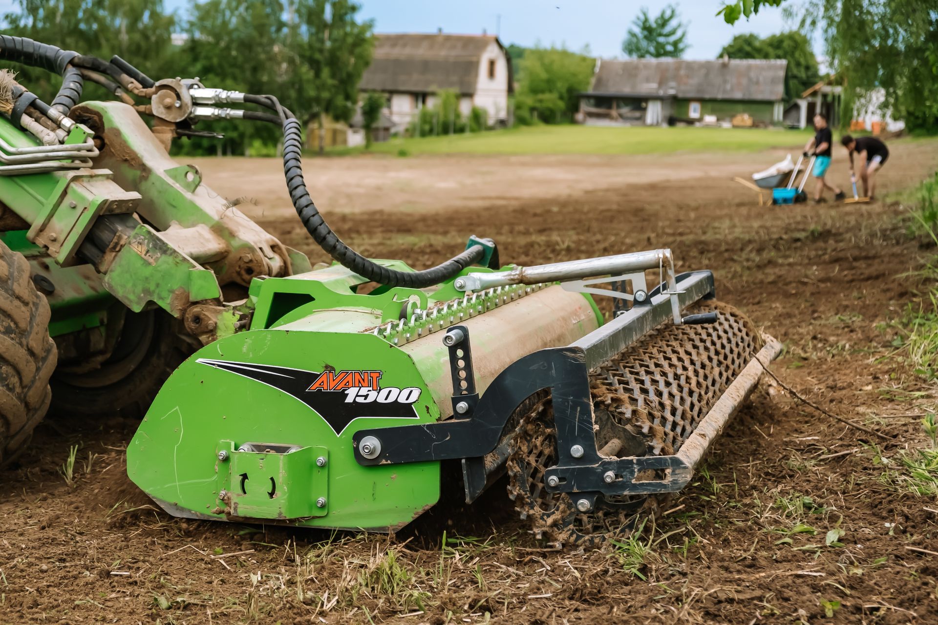 Green tractor with soil tiller working in a muddy field, with a few people in the background.