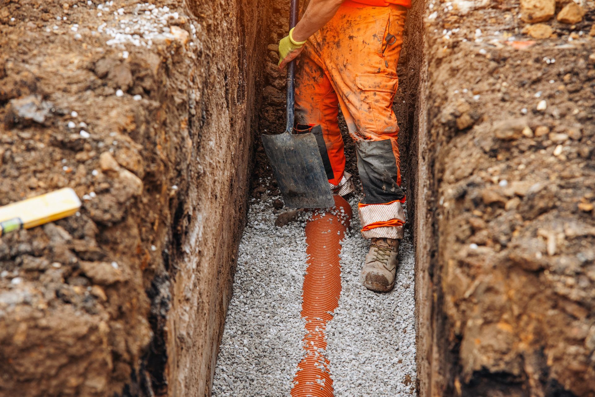 Worker shoveling wet concrete into a narrow trench with gravel base