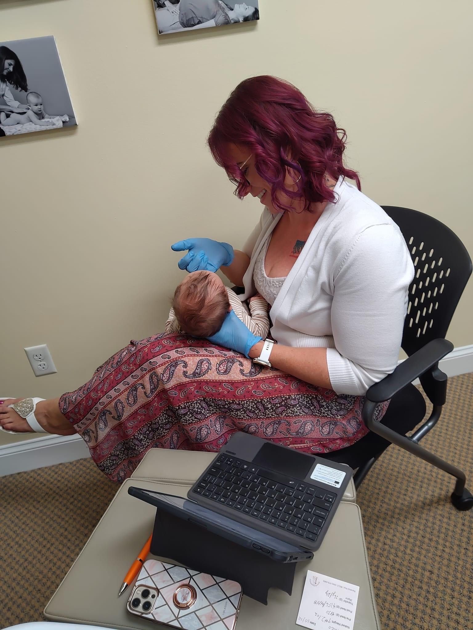 Person seated in a clinic chair holding a newborn, with a laptop on a nearby tray table.