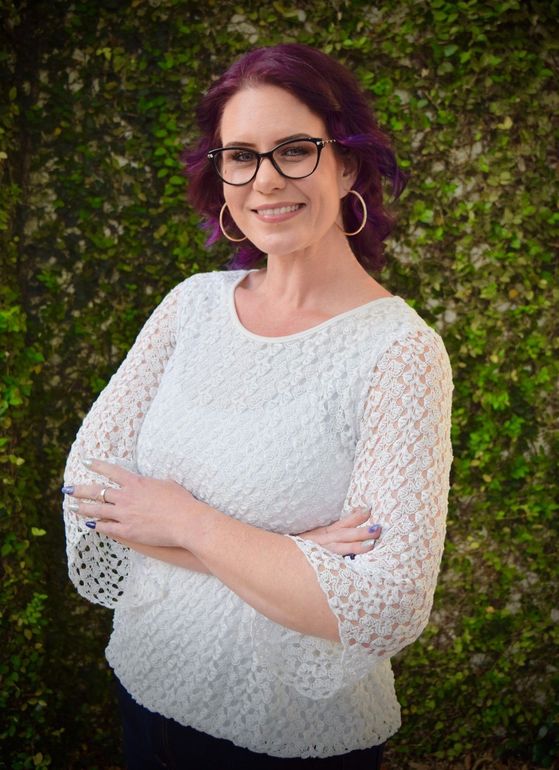 Woman with purple hair and glasses wearing a white lace top, standing with arms crossed against a leafy background