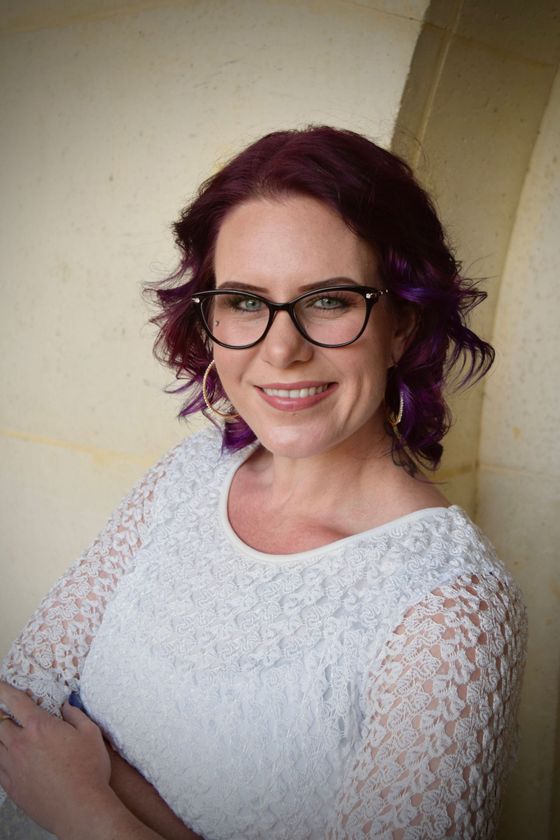 Woman with purple hair and glasses smiling in a white lace top against a beige wall