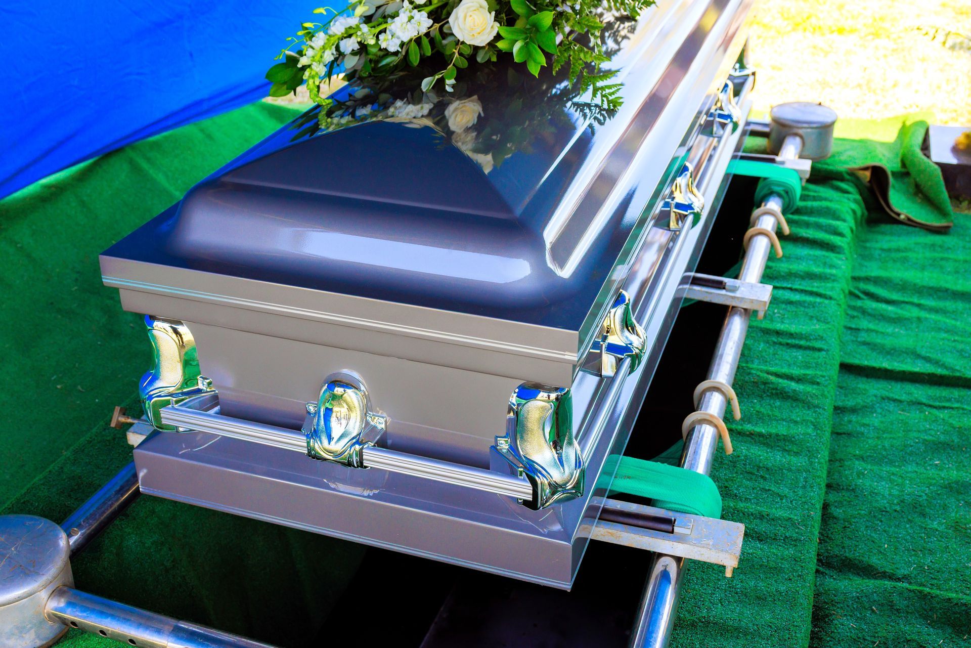 Silver casket with white flowers and blue ribbon on a green lawn, viewed at an angle