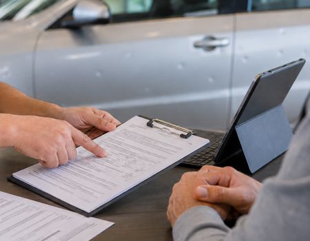 Two people review a form on a table beside a parked silver car, one pointing at the paperwork.