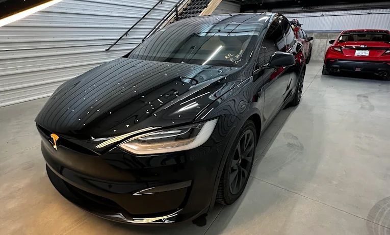 Black sports sedan in a garage, front view, with a red car parked behind it.